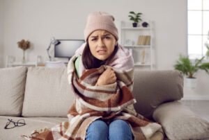 A woman sitting on a couch looking cold with a blanket around her.