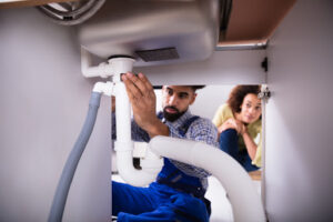 Rite Way of Phoenix plumber checking under sink to estimate drain cleaning service cost while homeowner looks behind him.
