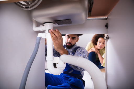 Rite Way of Phoenix plumber checking under sink to estimate drain cleaning service cost while homeowner looks behind him.