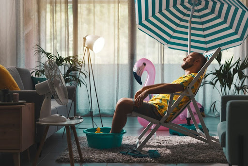 A man relaxing indoors with a beach chair, umbrella, and fan.