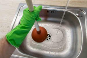 A close-up of a gloved hand using a plunger in a sink.